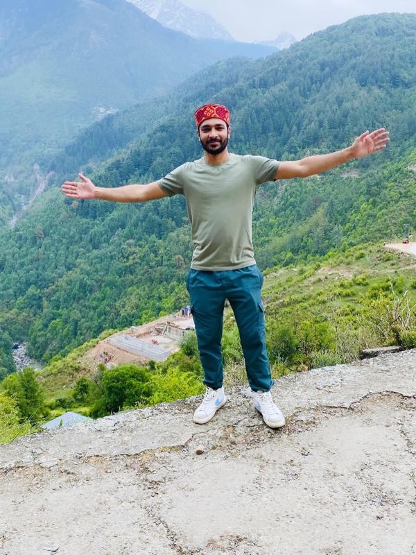 Saurav with his arms open on a ledge above a green, wooded valley