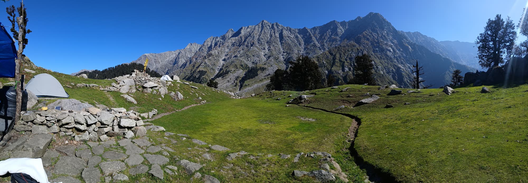A wide green meadow with tents and stone walls, below a long grey Himalayan ridge
